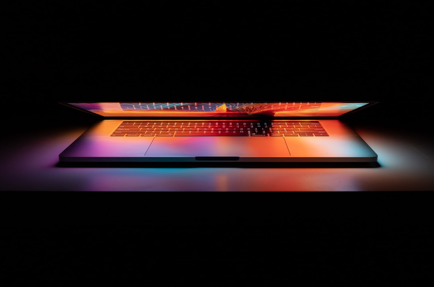 Half open laptop in a dark room. The colors are reflecting off the keyboard and white table under it.